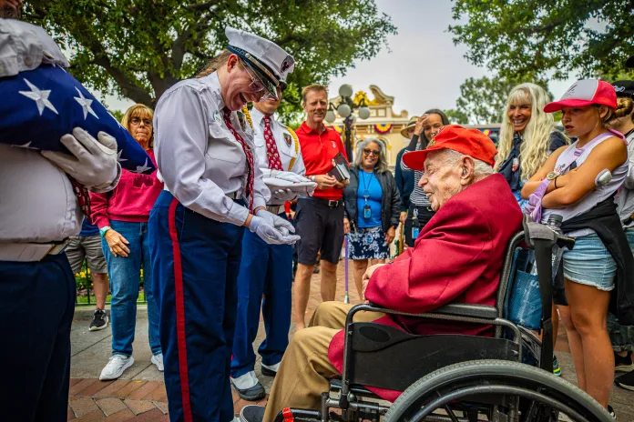 Anaheim, Calif. (March 31, 2026) – Disneyland Security Cast Member presents a challenge coin to 100-year-old World War II Veteran Charles Cram during the daily Flag Retreat ceremony at Disneyland Park where he was honored for his military service in front of park guests and his family. Cram served as a U.S. Navy Pharmacist’s Mate Second Class with the 5th Marine Division during the Battle of Iwo Jima — one of the most pivotal battles of the Pacific campaign. (Photo credit: Disney)