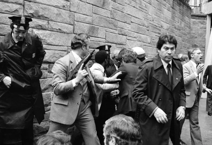 Secret Service agents and police officers swarm a gunman, obscured from view, after he attempted an assassination on President Ronald Reagan outside the Washington Hilton hotel