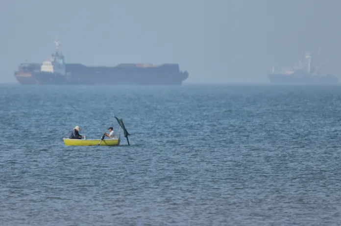 Two men sit in a small boat on the water as cargo ships are anchored in the background in the Strait of Hormuz off Bandar Abbas, Iran, Monday, May 4, 2026.