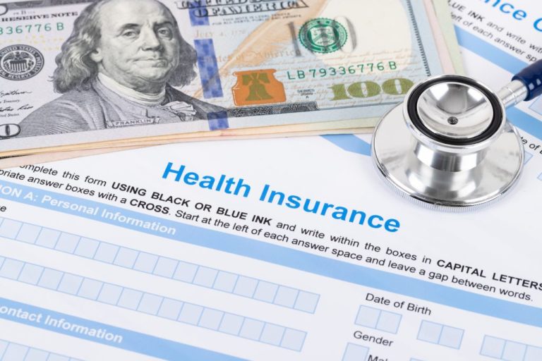 Health insurance papers with a $100 bill stacked atop and a stethoscope on the right. (Getty Images)