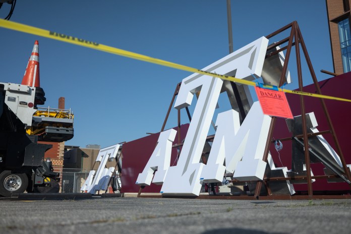 Large white "ATM" letters are being moved or installed outdoors, surrounded by construction equipment and yellow caution tape.