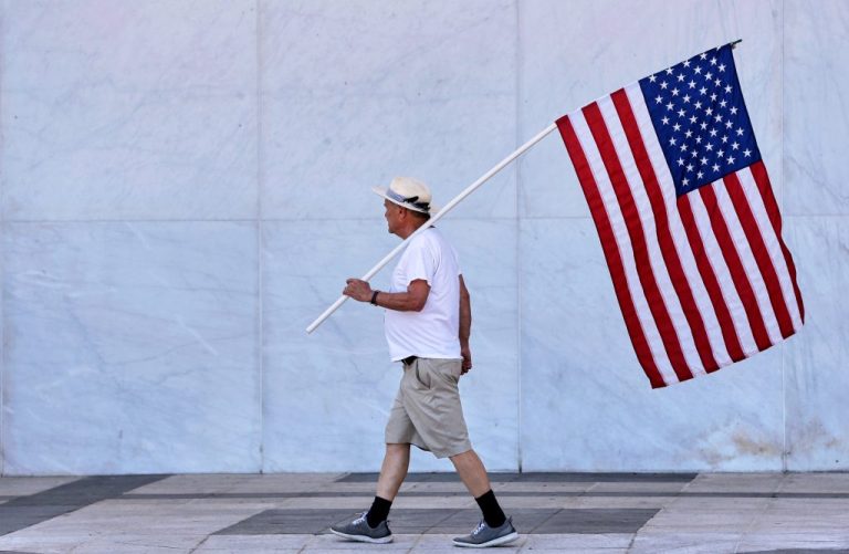 A man holds American flag as the wall of the John F. Kennedy Center for the Performing Arts is covered in tarp after President Donald Trump's name was removed, in Washington, Saturday, June 13, 2026. (AP Photo/Rahmat Gul)