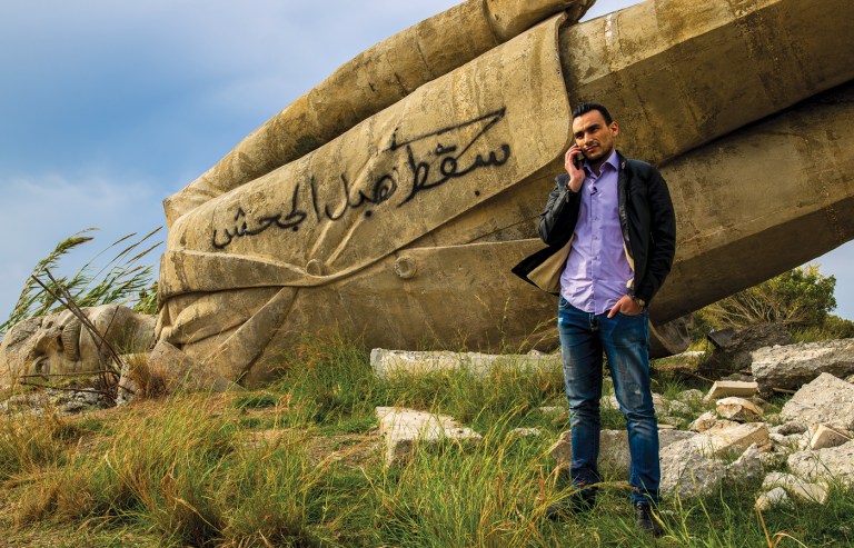 Hassan Ahmad, 27, an activist who works with NGOs and foreign journalists, stands beside the remains of a toppled Hafez al-Assad statue marked with graffiti reading, “The executioner’s rule has fallen.” (Photo by Jordan Allott)