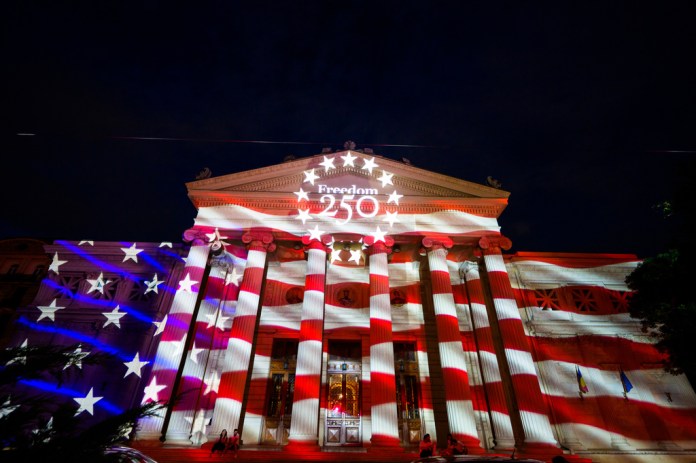 People sit on the steps of the Romanian Athenaeum, a 19th century built concert hall, as the U.S. flag is projected on it's facade ahead of United States Independence Day, in Bucharest, Romania, Friday, July 3, 2026. 