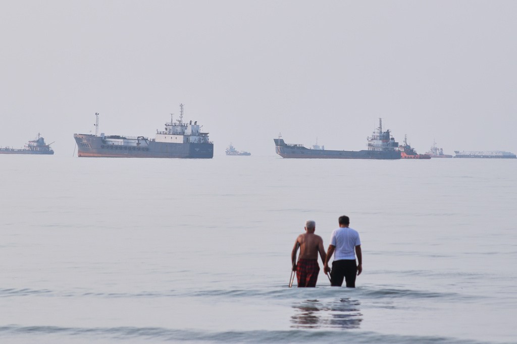Two men wade in the waters of the Strait of Hormuz with vessels anchored.