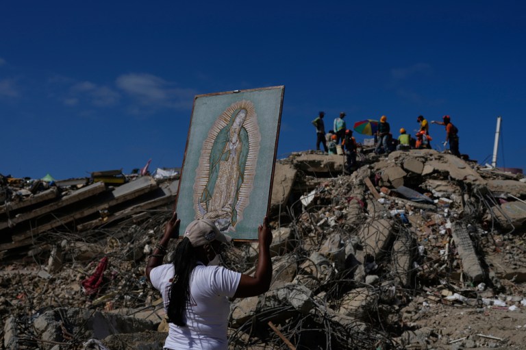 A woman holds a painting of the Virgin Mary as she accompanies a Catholic priest supporting people searching for the remains of relatives in the rubble of residential buildings that collapsed during the earthquakes that struck La Guaira, Venezuela, Friday, July 17, 2026.