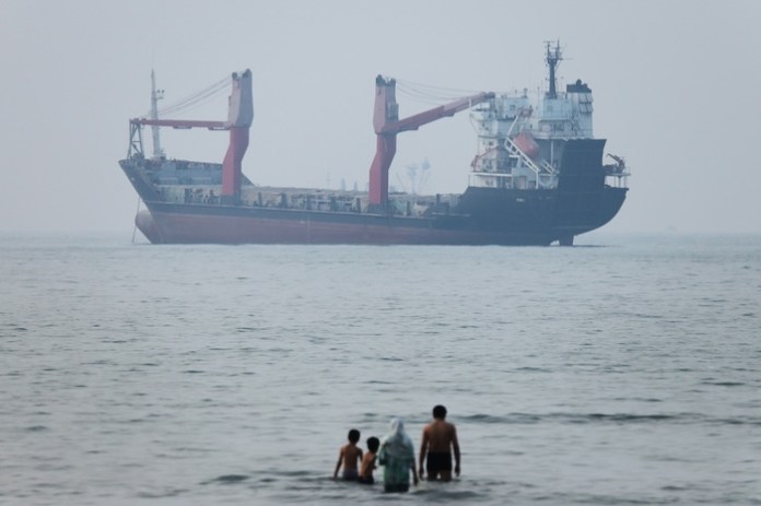 A group of people stands in shallow water as a cargo ship appears anchored in the Strait of Hormuz off Bandar Abbas, Iran, Tuesday, June 30, 2026. (Amirhosein Khorgooi/ISNA via AP)