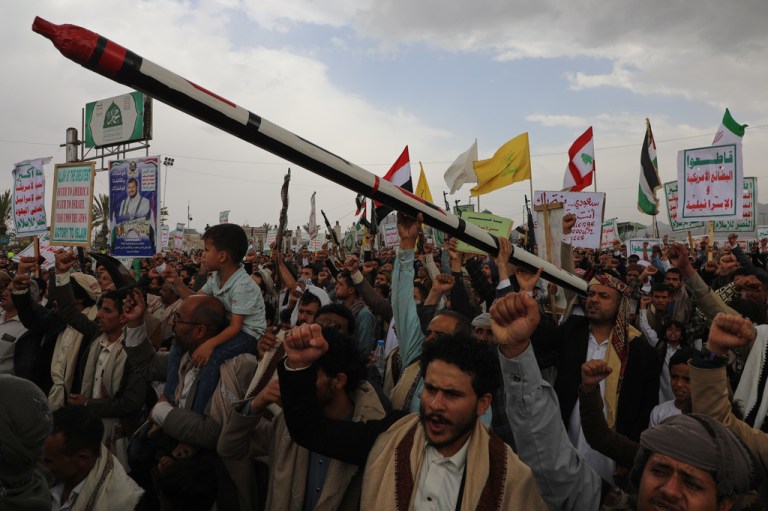 Thousands crowd around adorning flags for a Houthi parade. A kid is seen on an adult's shoulders and many have their fists raised in the air.
