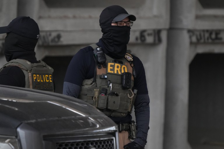 An Immigration and Customs Enforcement officer concealed by a black cloth facial covering walks the scene where a man was shot in his vehicle by an ICE officer.