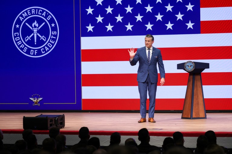 Secretary of Defense Pete Hegseth waves as he gives a State of the Force address onstage at Marine Corps Base Quantico against a backdrop of a huge American flag.