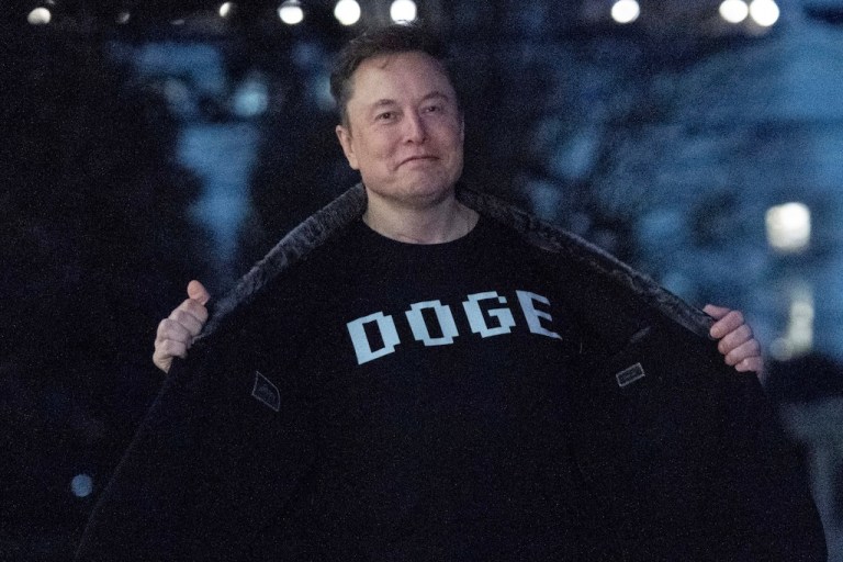 Elon Musk flashes his T-shirt that reads "DOGE" to the media as he walks on South Lawn of the White House, in Washington, Sunday, March 9, 2025.