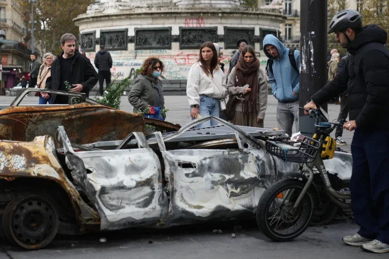 People watch a burned-out car after a quiet night under curfew